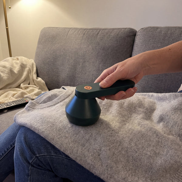Green electric lint remover used on a grey jumper on a sofa