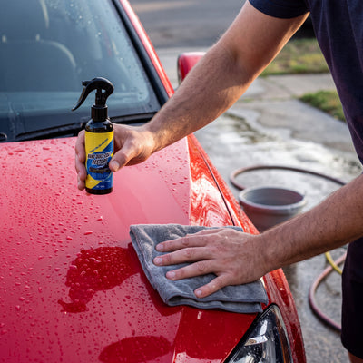 A hand wiping the wet hood of a red car with a grey microfiber cloth while the other hand holds the Novum Car Nano Coat Spray bottle