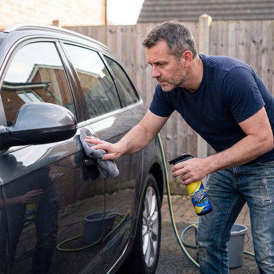 A man wiping the door of a dark grey car with a microfiber cloth, the Novum Car Nano Coat Spray bottle in his other hand