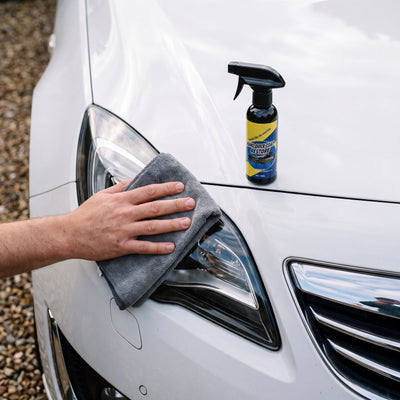 A hand wiping a car headlight with a grey microfiber cloth, the Novum Car Nano Coat Spray bottle standing on the white hood